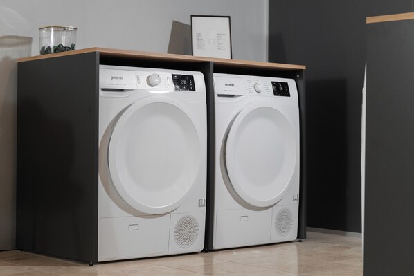 Two white front-loading washing machines with control panels, placed on a dark floor against a wall with a framed picture above. - master Linzergasse (Salzburg)
