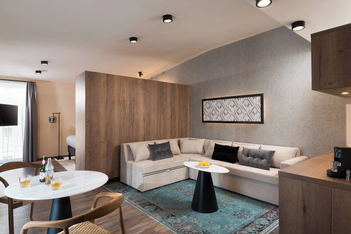 A modern living room with a beige sectional sofa, a round dining table, and a coffee machine on a wooden counter.