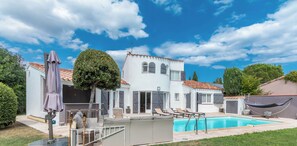 Uma casa de dois andares com piscina, móveis de exterior e uma rede.