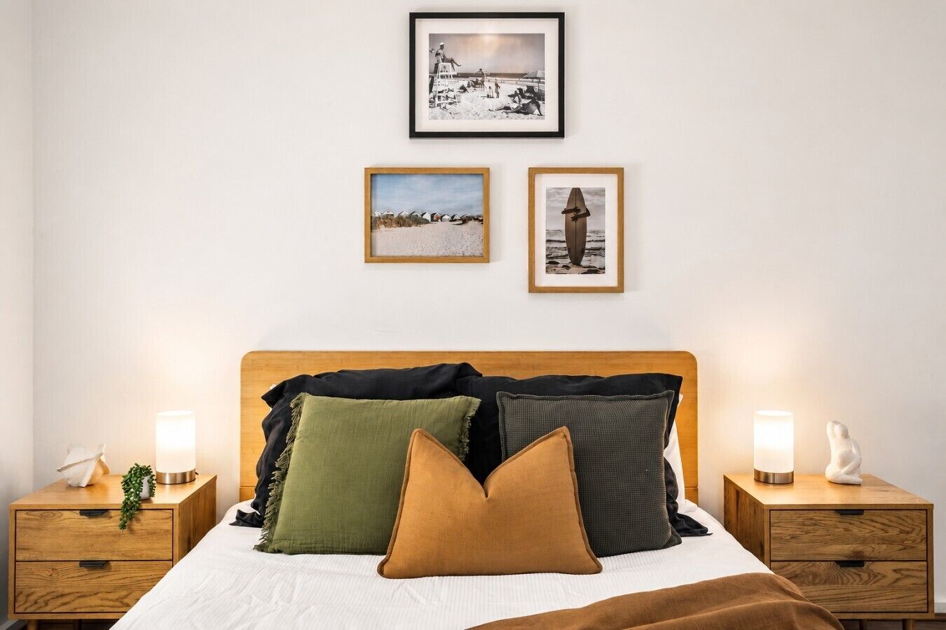 A bedroom with a wooden headboard, a bed with white bedding, and three pillows in different colors. There are two bedside tables with lamps and a small statue on one.