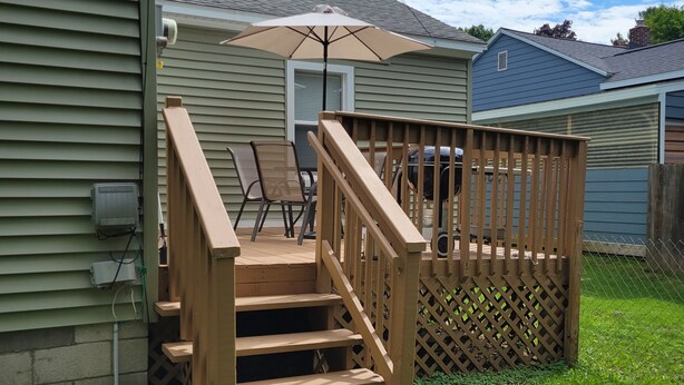 A wooden deck with a railing, a patio table, and a chair, adjacent to a house with a garage. - Dog-Friendly, Fenced-In Backyard, Quiet Neighborhood (Sturgeon Bay)
