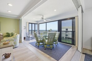 Quarto espaçoso com área de jantar, ventilador de teto e grandes janelas com vista para o mar.