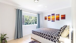 Een slaapkamer met een bed, een raam met gordijnen, een kamerplant en vier schilderijen van een zonsondergang aan de muur.