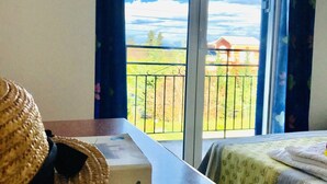A room with a view of a green landscape through a sliding door, a wooden table with a box and a straw hat on it, and a bed with a striped bedspread. - Banos apartments and swimming  pool (Corfu)