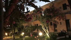 A courtyard with a building, trees, and pink flowers. - Banos apartments and swimming  pool (Corfu)