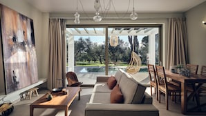 A modern living room with a sofa, coffee table, and dining area. There is a large window with a view of a pool and trees. - Metis Villa, a Luxury Retreat (Zakynthos)