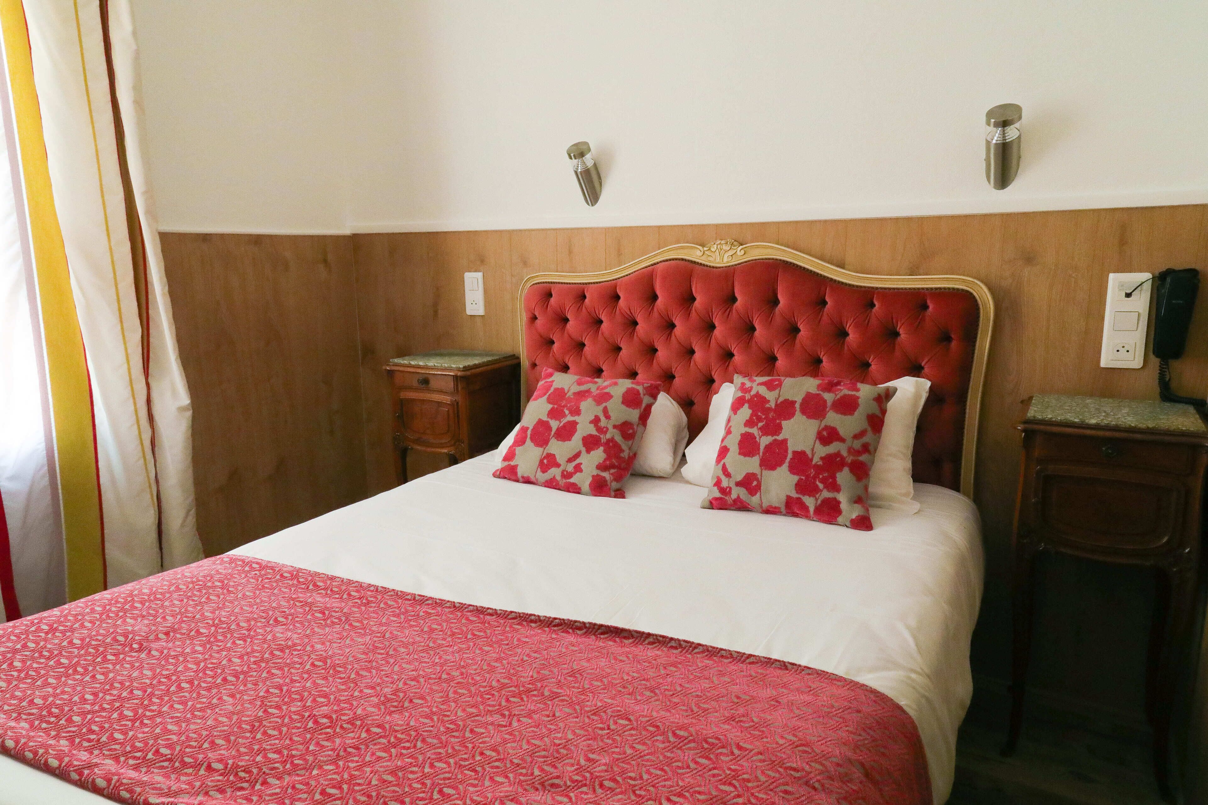 A bed with a red headboard, white bedding, and floral pillows. There are two bedside tables, one with a lamp and the other with a phone.