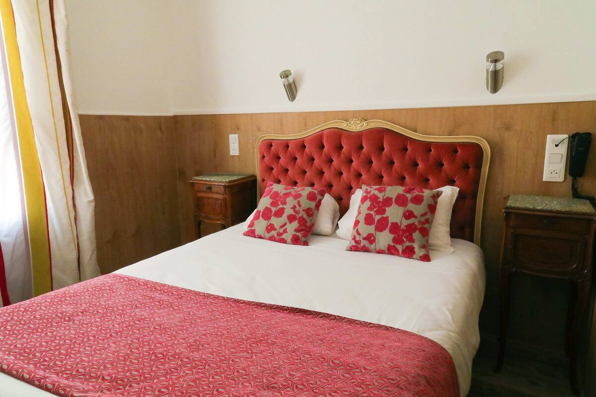 Una cama con cabecera roja, ropa de cama blanca y almohadas florales. Hay dos mesitas de noche, una con una lámpara y la otra con un teléfono.