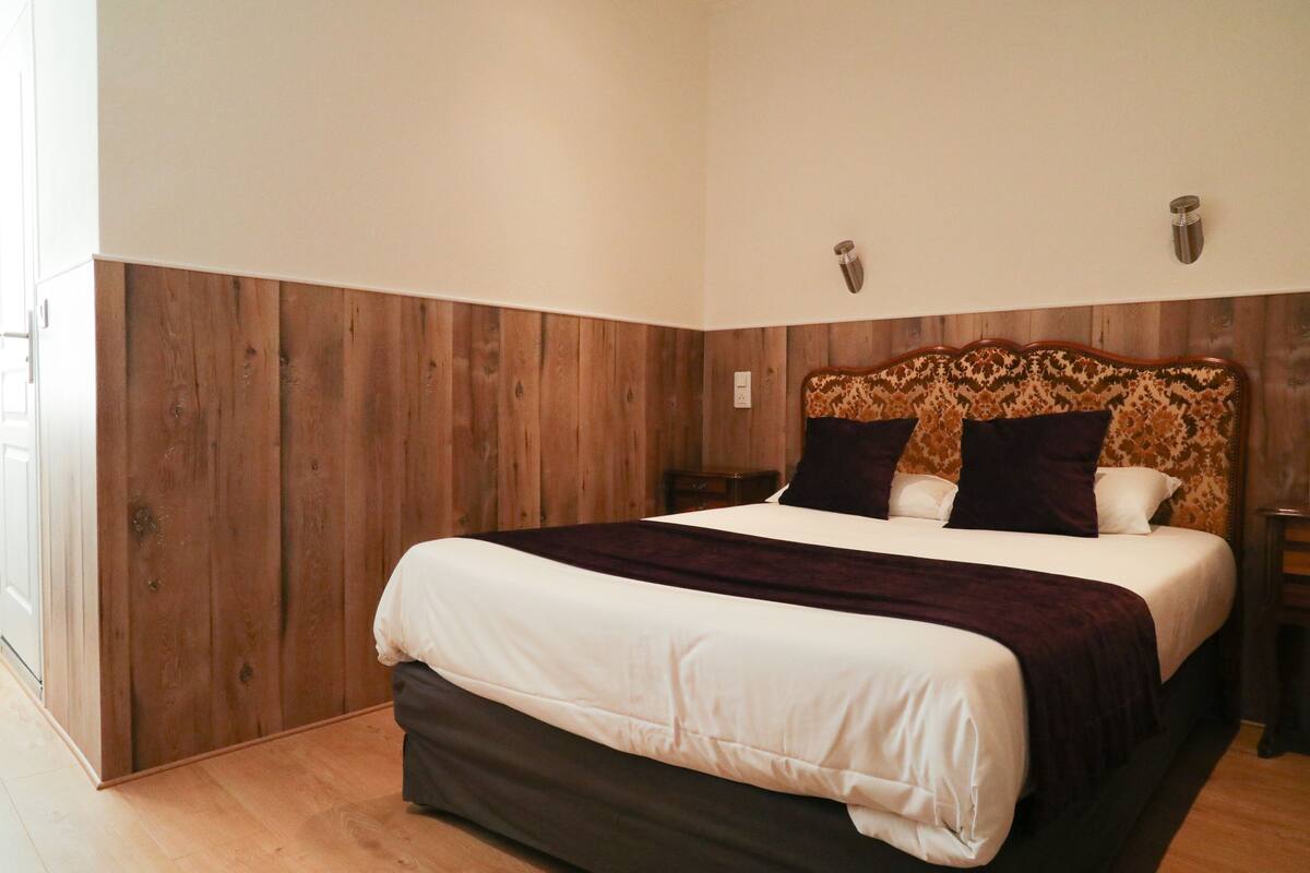Un dormitorio con cabecera de madera, una cama grande con ropa de cama blanca y oscura, y una mesita de noche con una lámpara.