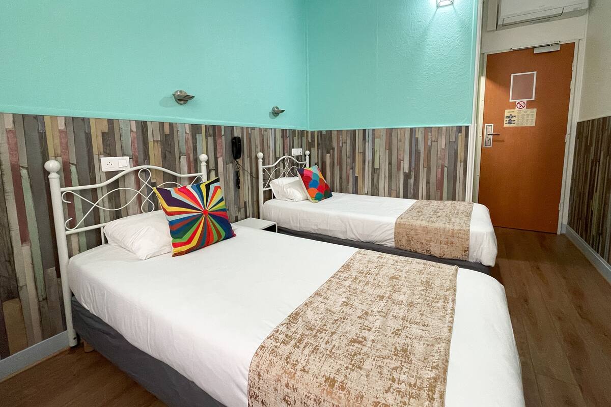 Habitación de hotel con dos camas, cada una con colchón blanco y almohada de colores. La habitación tiene una pared verde azulado y un detalle en panel de madera.