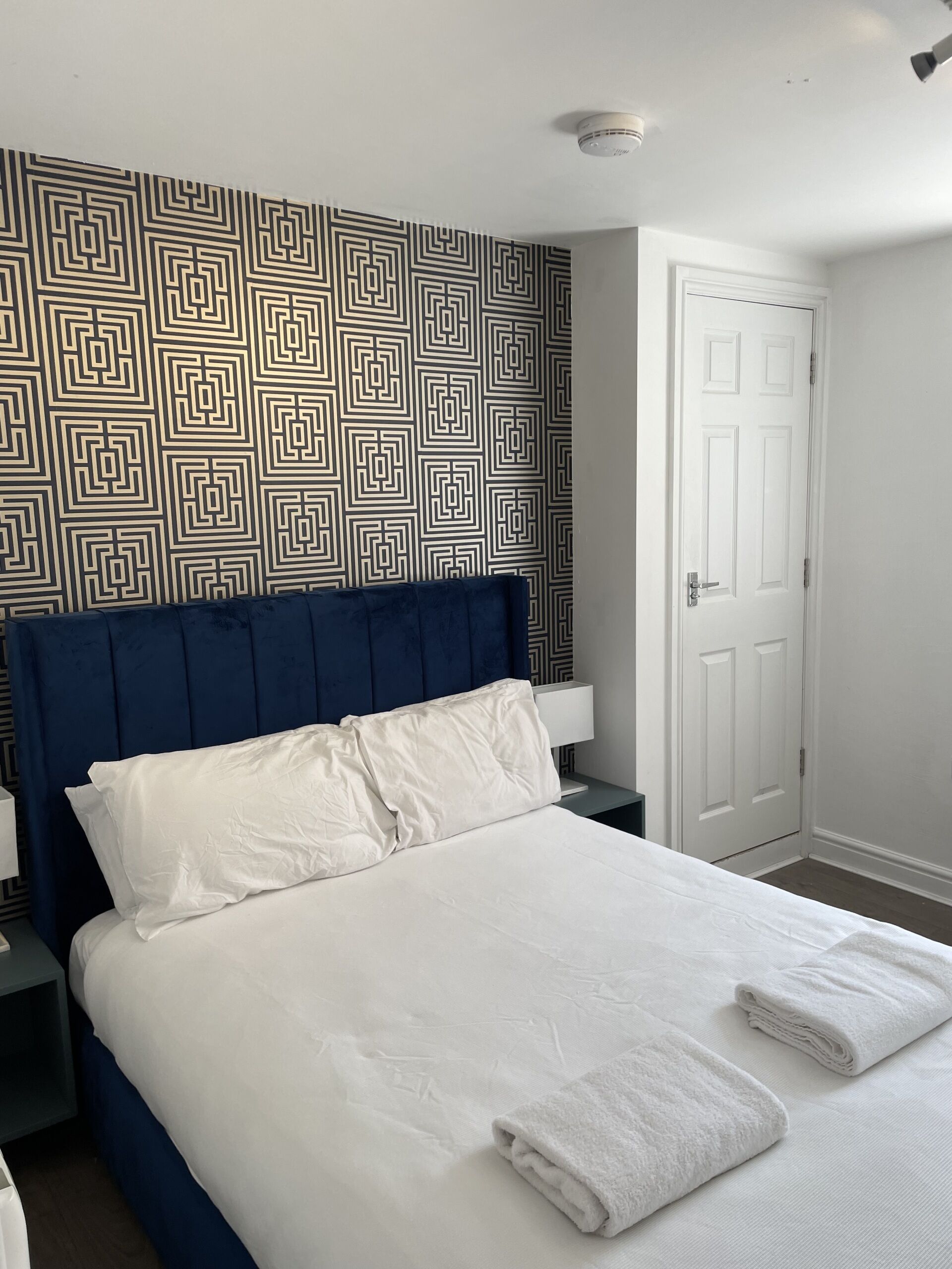 A neatly made bed with white linens and two folded towels, a dark blue headboard, a patterned wall, a white door, and a nightstand with a lamp.