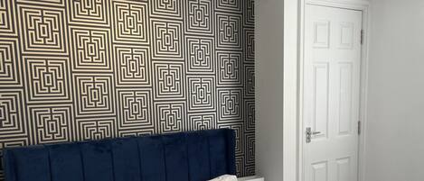 A neatly made bed with white linens and two folded towels, a dark blue headboard, a patterned wall, a white door, and a nightstand with a lamp.
