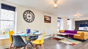 A modern living room with a dining area, featuring a large wall clock, a sofa, and a dining table with chairs. - Whitby Bridge Luxury Apartments (Whitby)