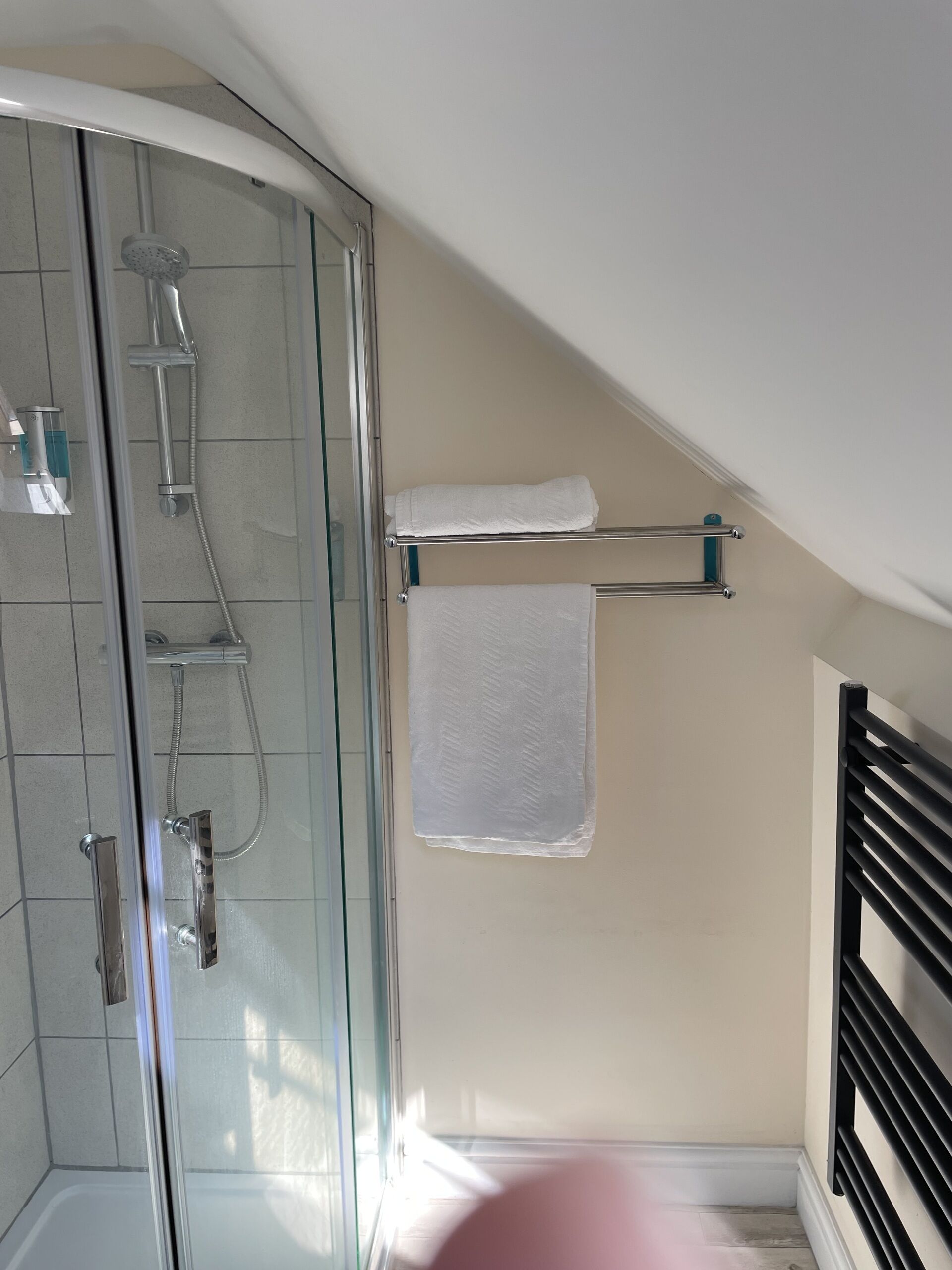 A glass-enclosed shower with a handheld showerhead and a handheld faucet. There is a towel rack with two towels hanging on it.
