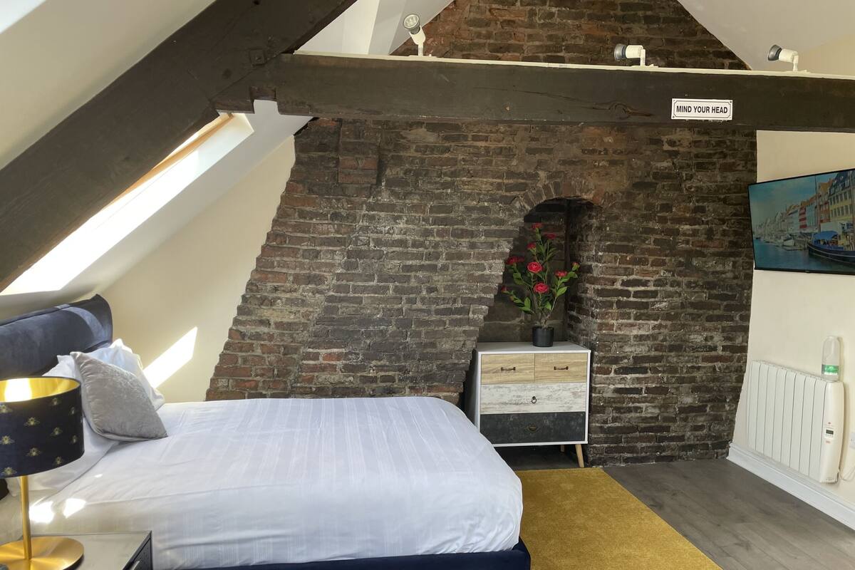 A bedroom with a sloped ceiling, a brick wall, a bed with white bedding, a nightstand with a potted plant, and a sign that says "MIND YOUR HEAD".