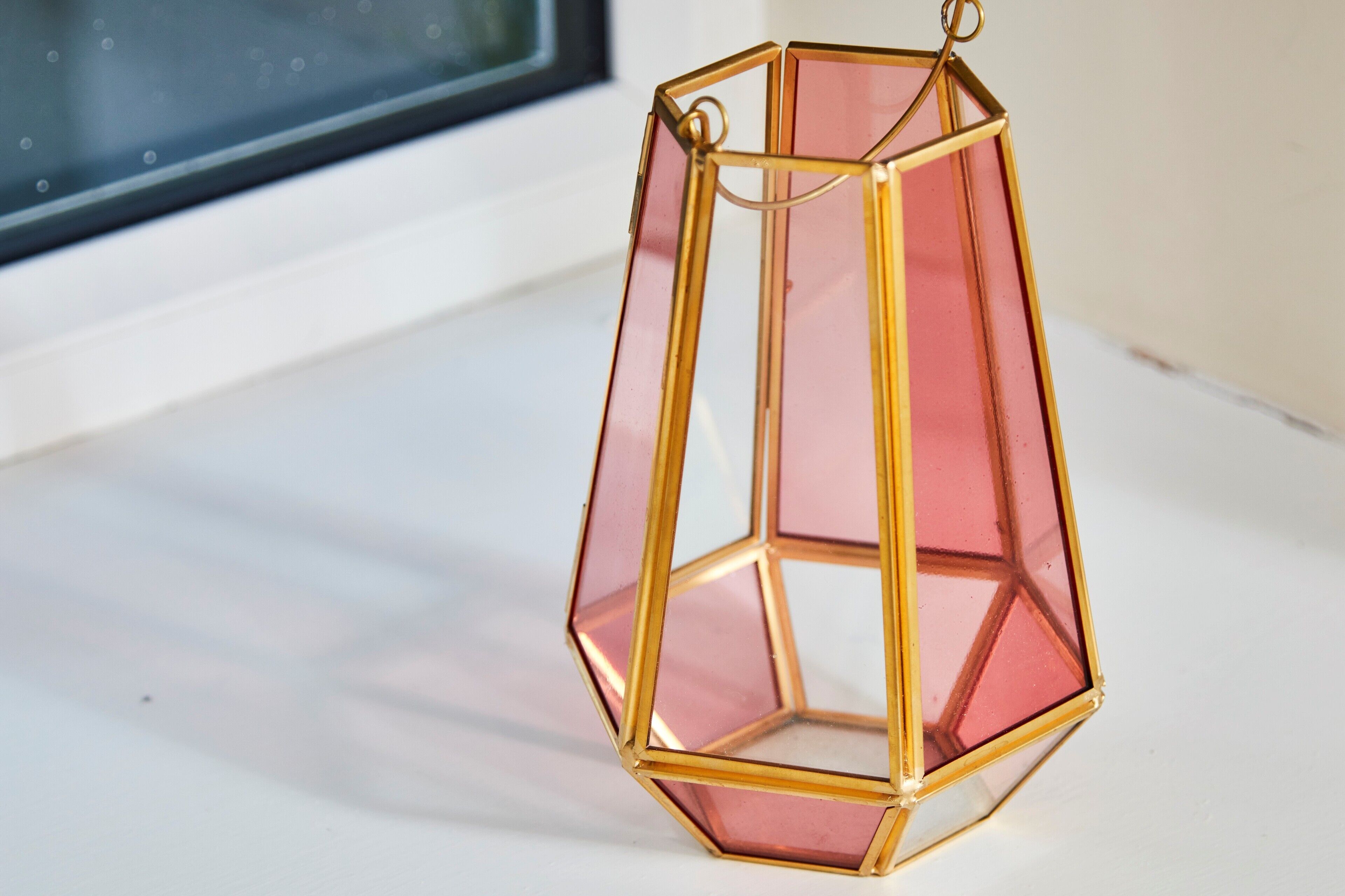 A hexagonal, gold-framed, pink-tinted lantern hanging by a window.