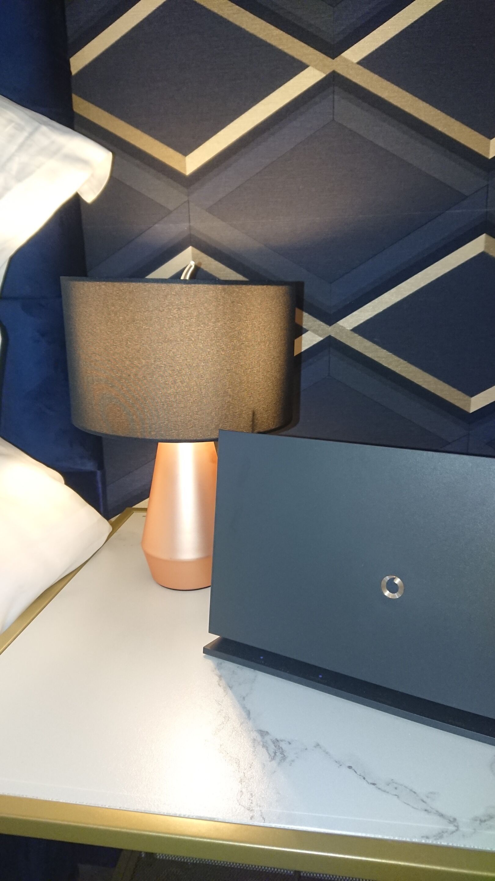 Een bed met een wit kussen, een laptop op een marmeren oppervlak, een lamp met een beige kap, en een wand met een geometrisch patroon.