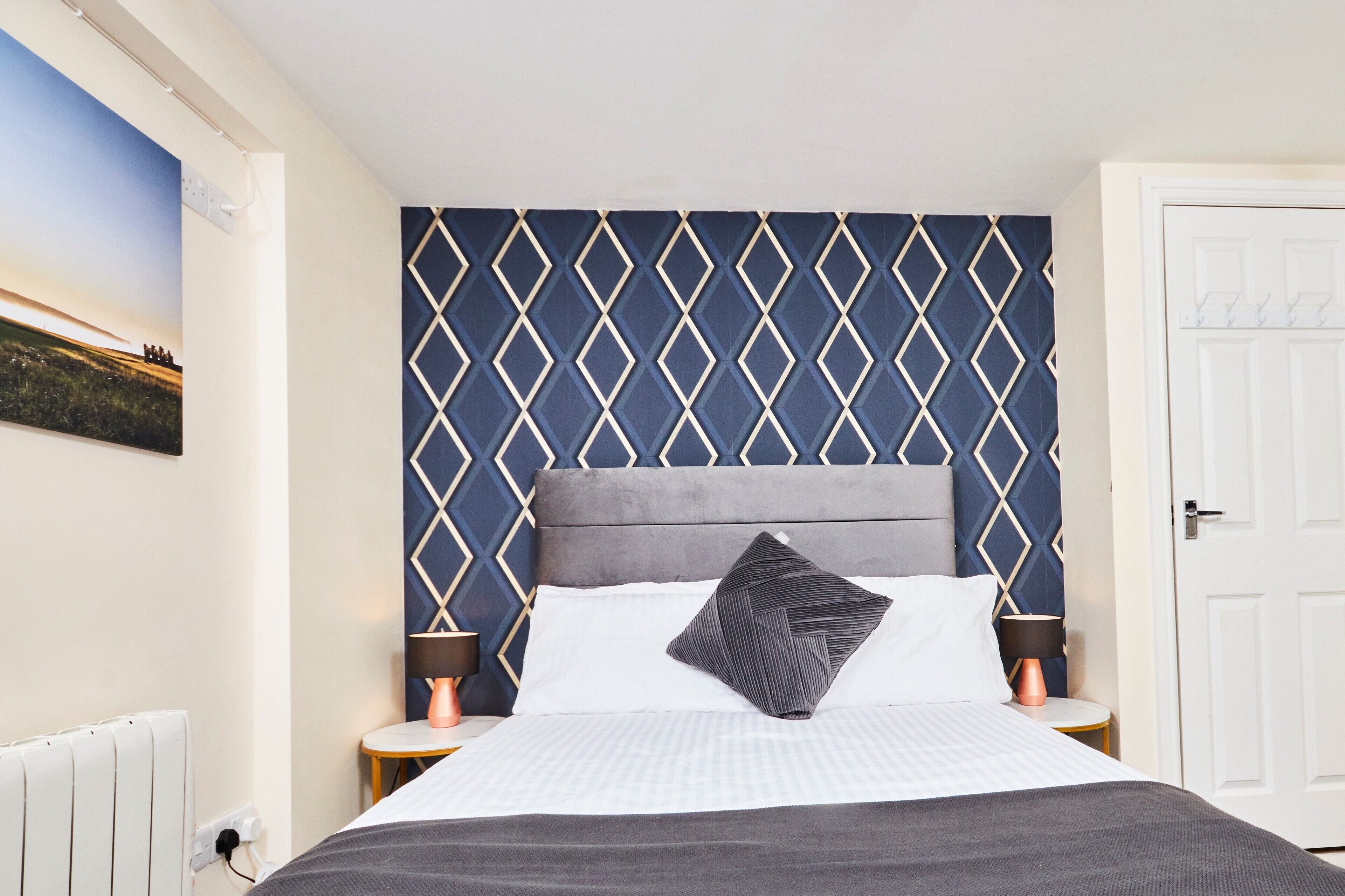 Un lit bien fait, avec une tête de lit grise, du linge de lit blanc et un seul oreiller noir. Deux tables de chevet avec des lampes sont placées de chaque côté du lit. Le mur derrière le lit est recouvert d’un papier peint à motifs géométriques.