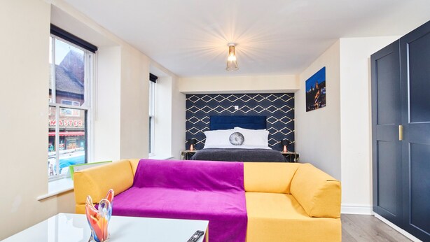 A hotel room with a bed, a yellow and purple sofa, a white table with a remote control, and a window with a view of a building. - Whitby Bridge Luxury Apartments (Whitby)