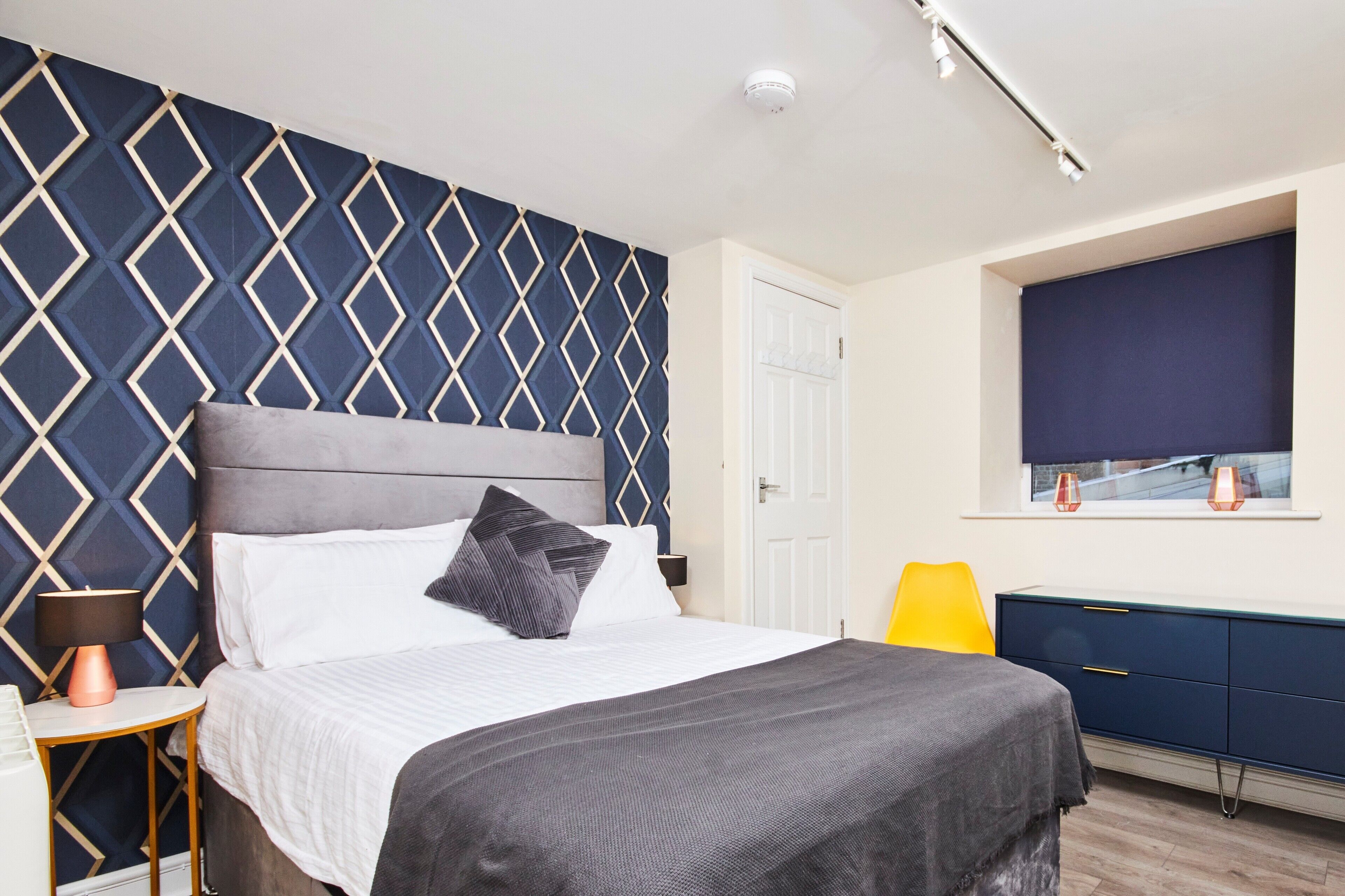 A modern bedroom with a geometric-patterned headboard, a bed with white linens and a grey throw, a bedside lamp, a yellow chair, and a window with a view.