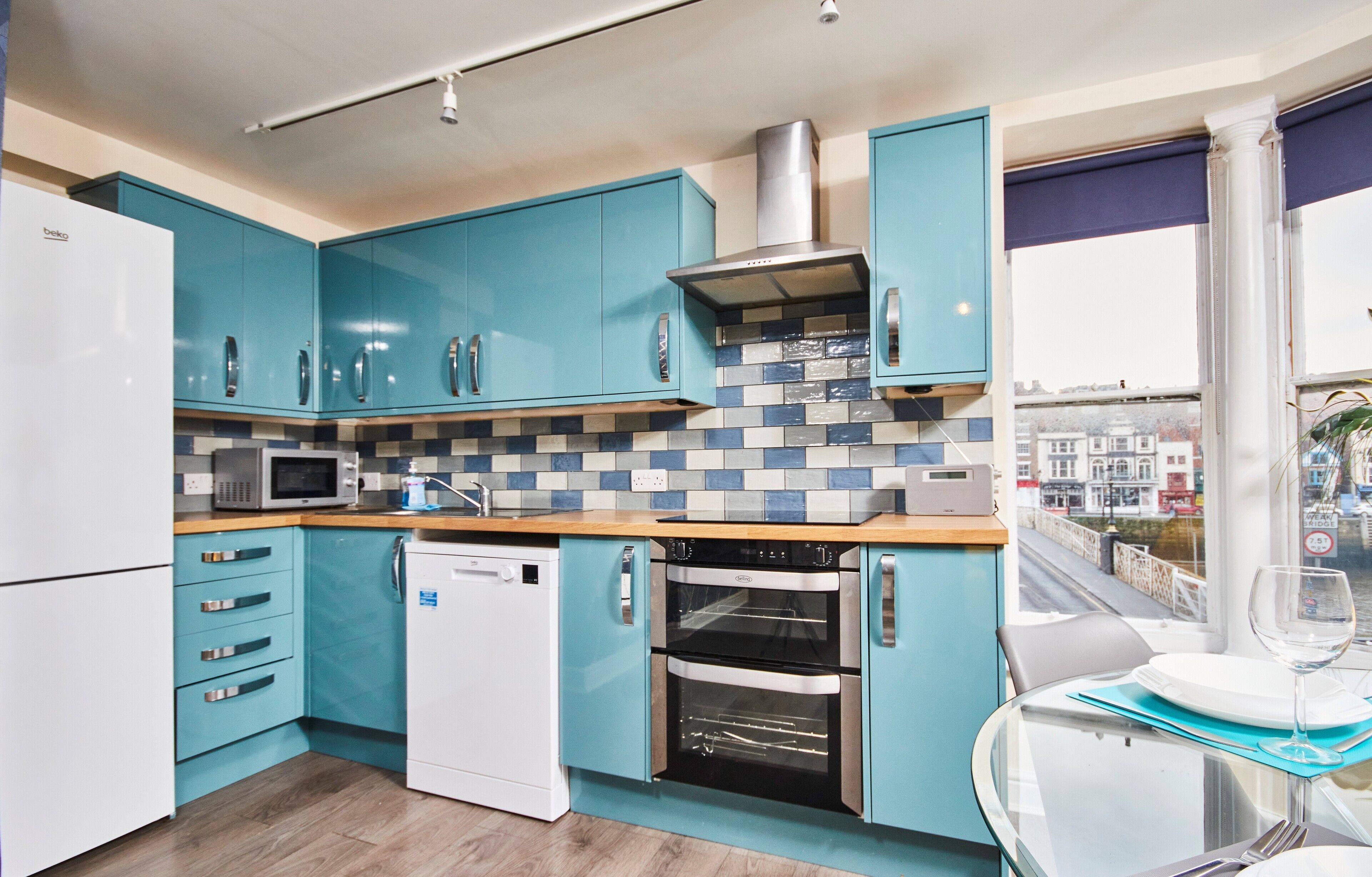 A modern kitchen with teal cabinets, a white refrigerator, a microwave, a dishwasher, and two ovens.