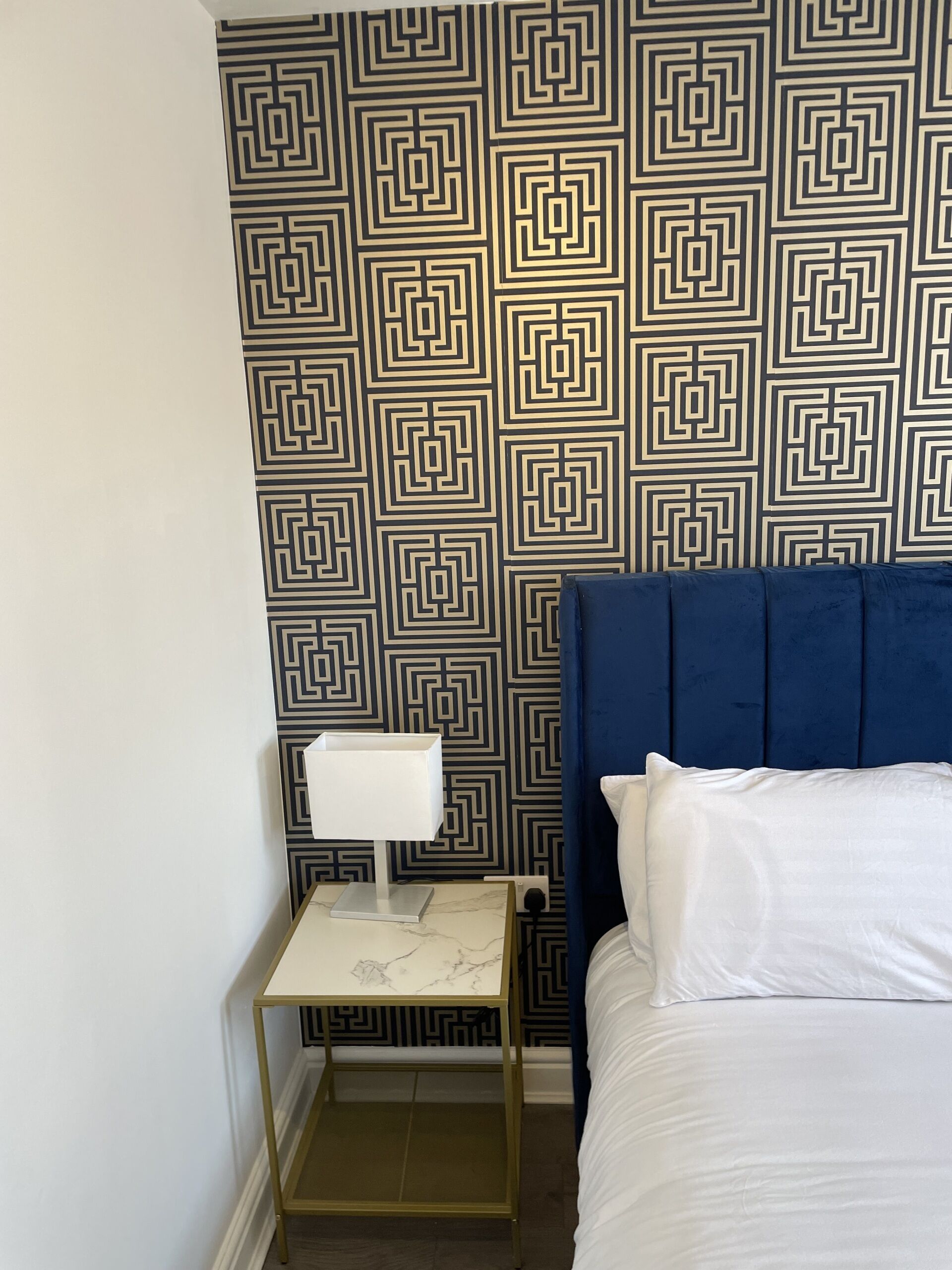 A bedroom with a patterned wallpaper, a blue headboard, a bedside table with a lamp, and a white bed.