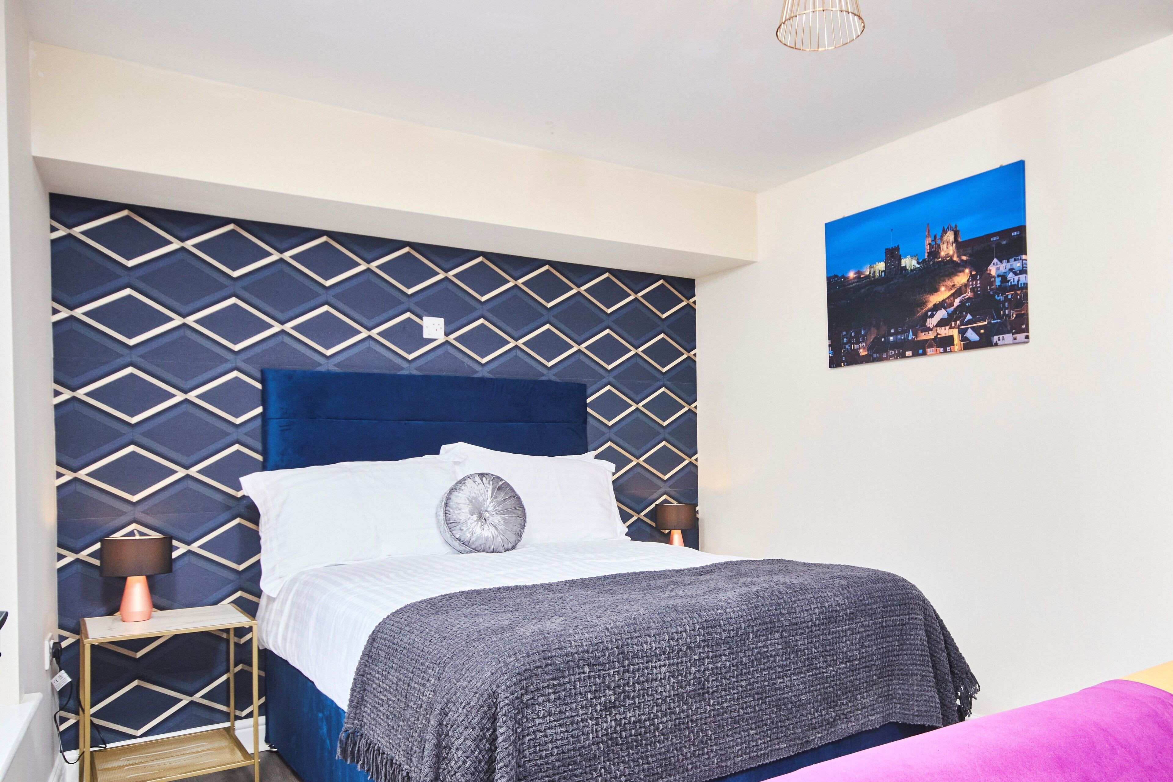 Une chambre avec un mur orné d’un motif géométrique, un grand lit avec une tête de lit bleue, un tableau représentant un paysage urbain et une table de chevet avec une lampe.