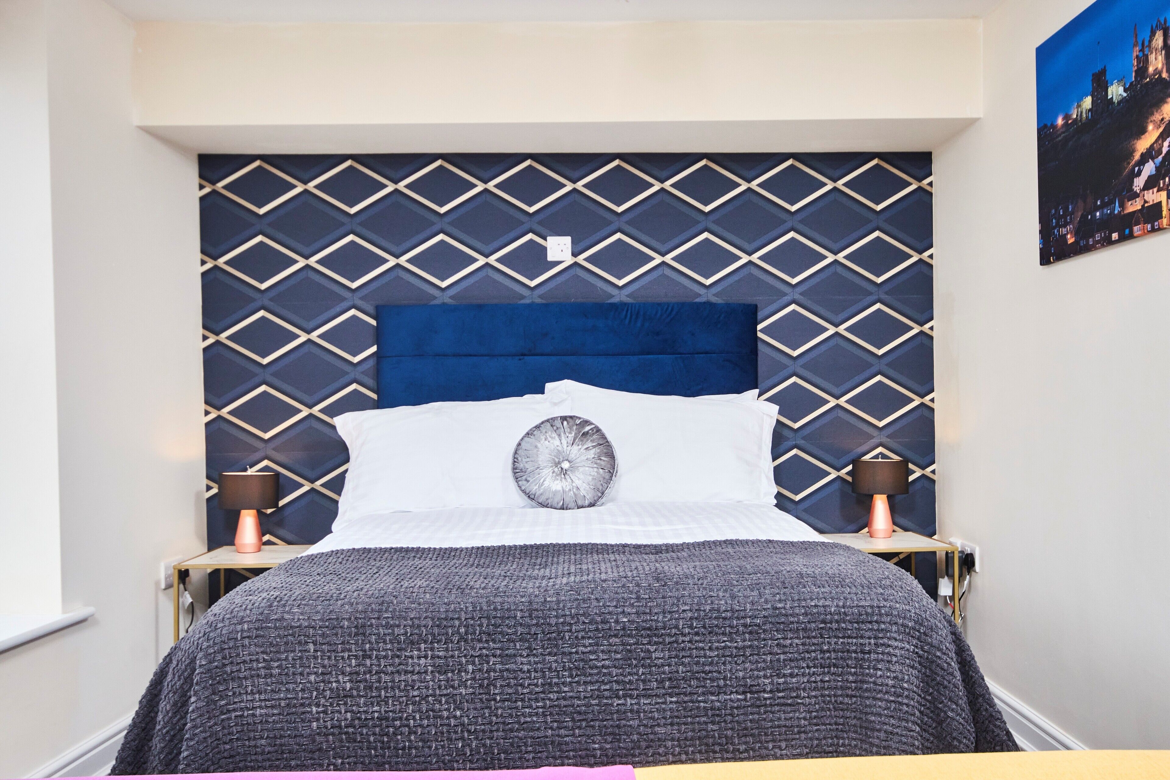 A neatly made bed with a blue headboard, two bedside lamps, and a wall-mounted picture.
