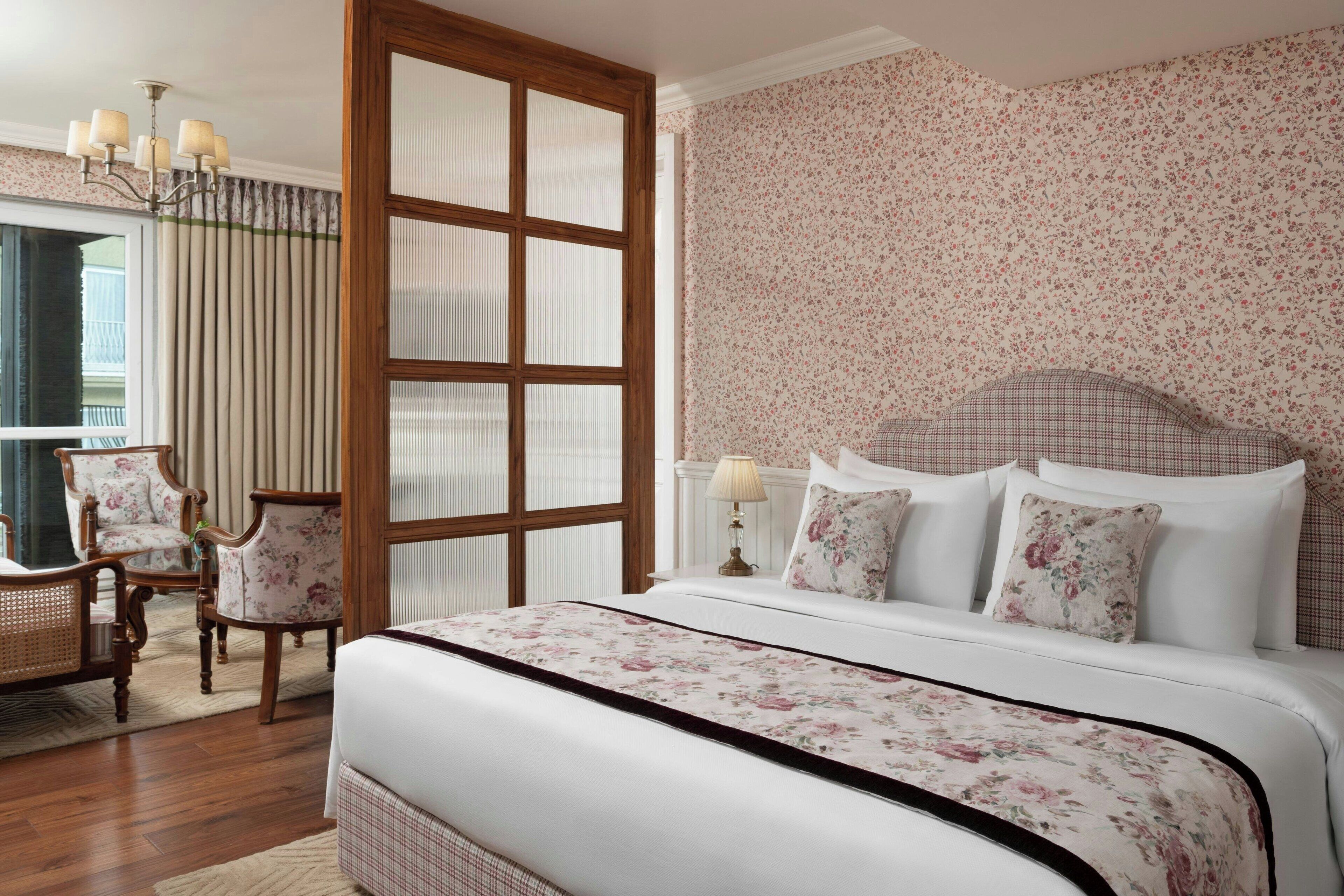 Une chambre à coucher avec un grand lit, un papier peint à motifs floraux, une fenêtre à cadre en bois et un lustre classique.