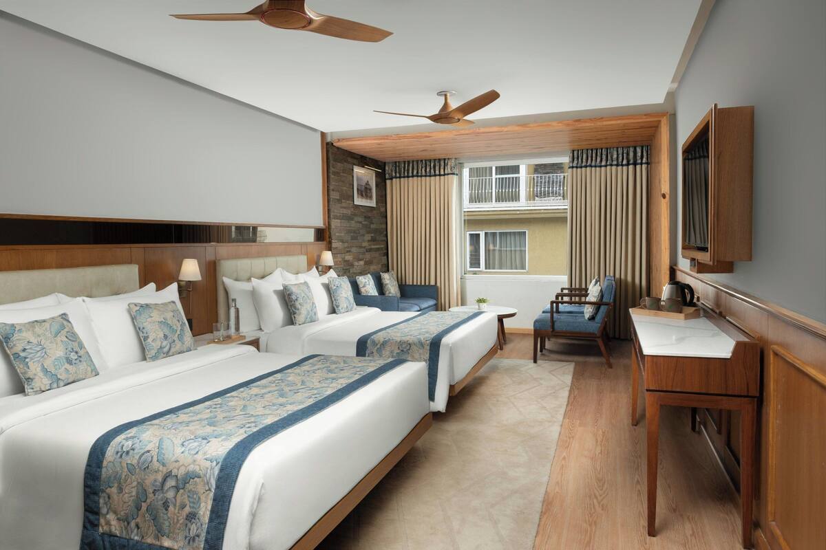 A hotel room with two beds, a desk, a chair, a ceiling fan, and a window with curtains.