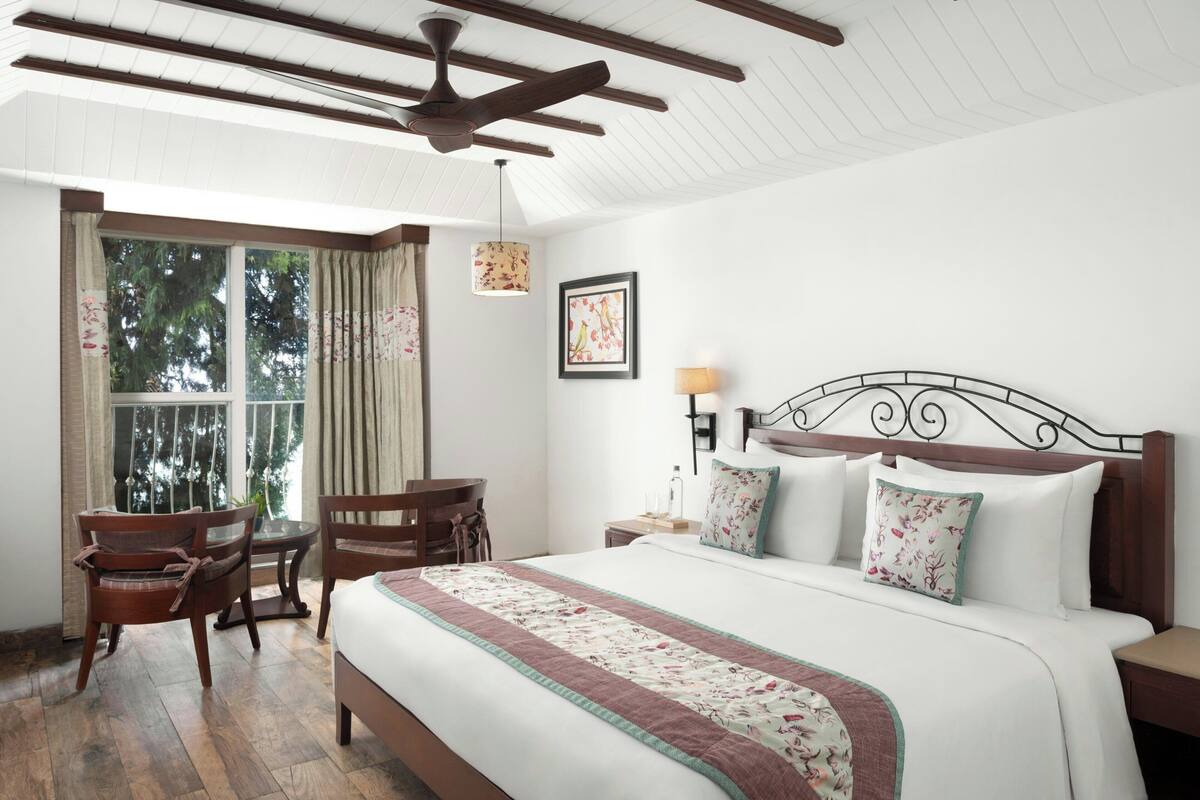 A hotel room with a large bed, wooden headboard, a desk with a chair, a window with curtains, and a ceiling fan.