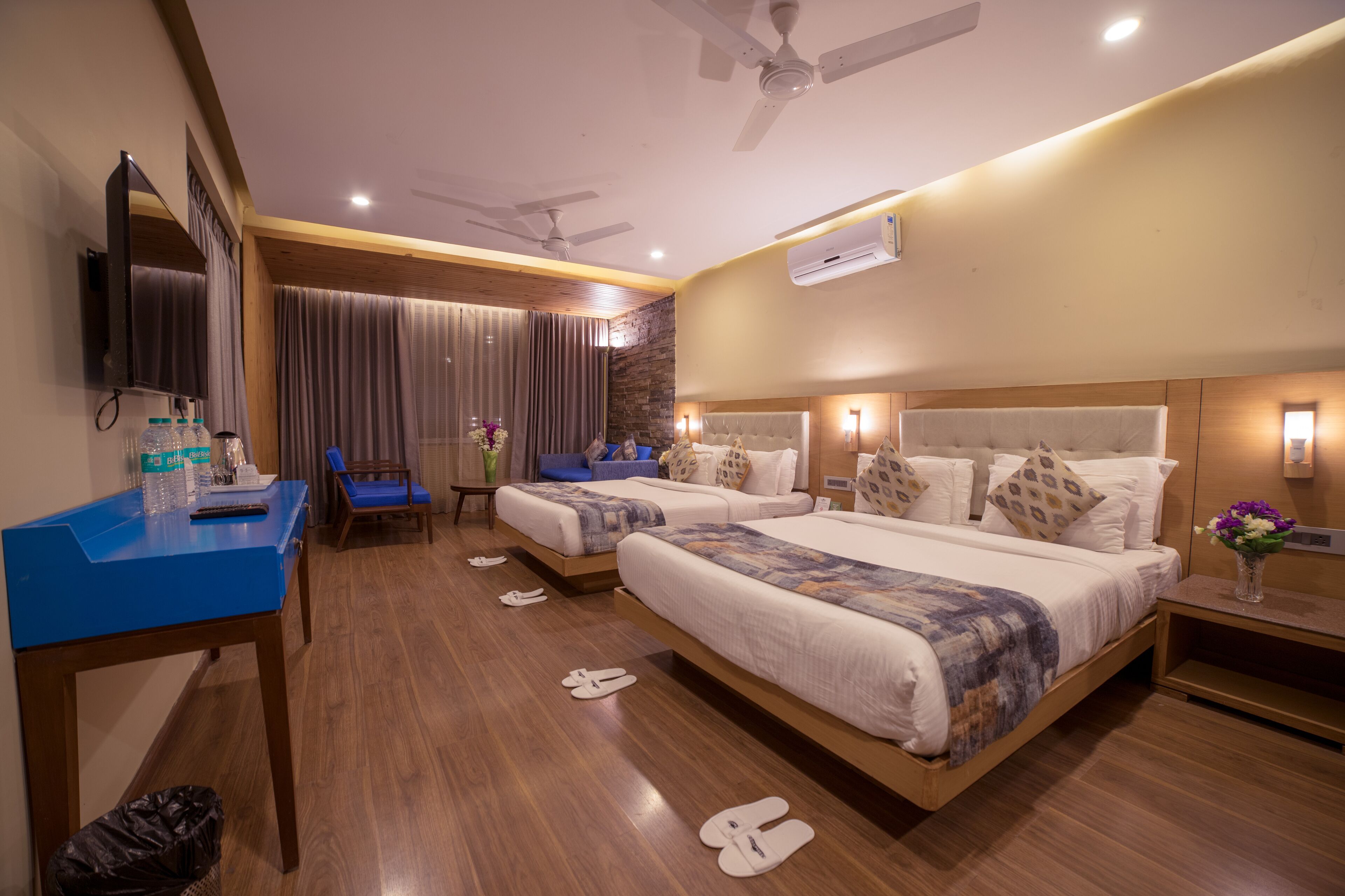 A hotel room with two beds, a desk, a chair, a TV, and an air conditioning unit.