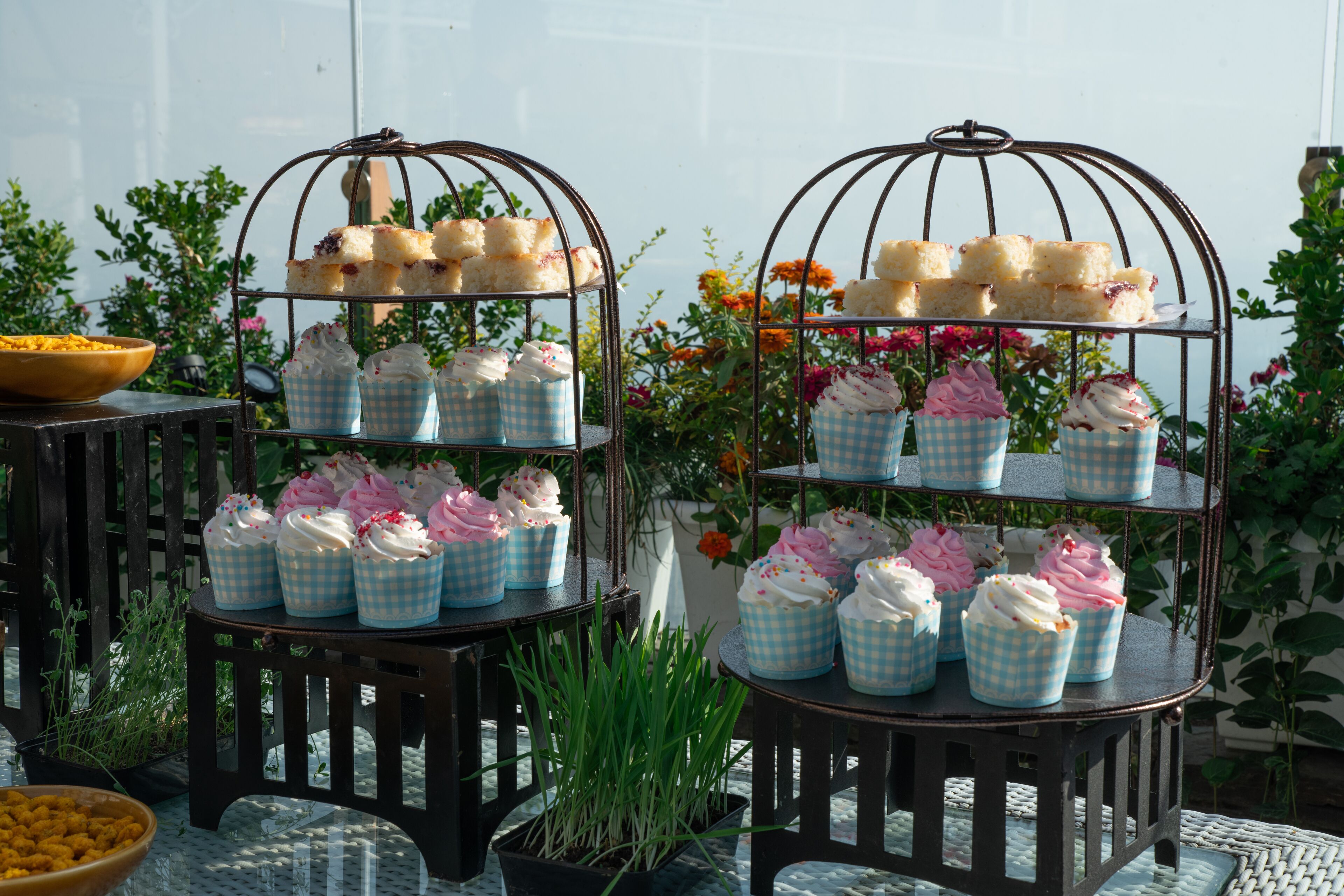 Two metal tiered stands with cupcakes and sandwiches, surrounded by potted plants and flowers.