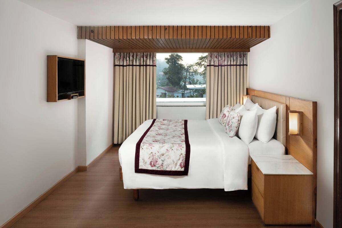 A hotel room with a wooden headboard, a bed with white bedding and a floral throw, a bedside table with a lamp, and a TV mounted on the wall.