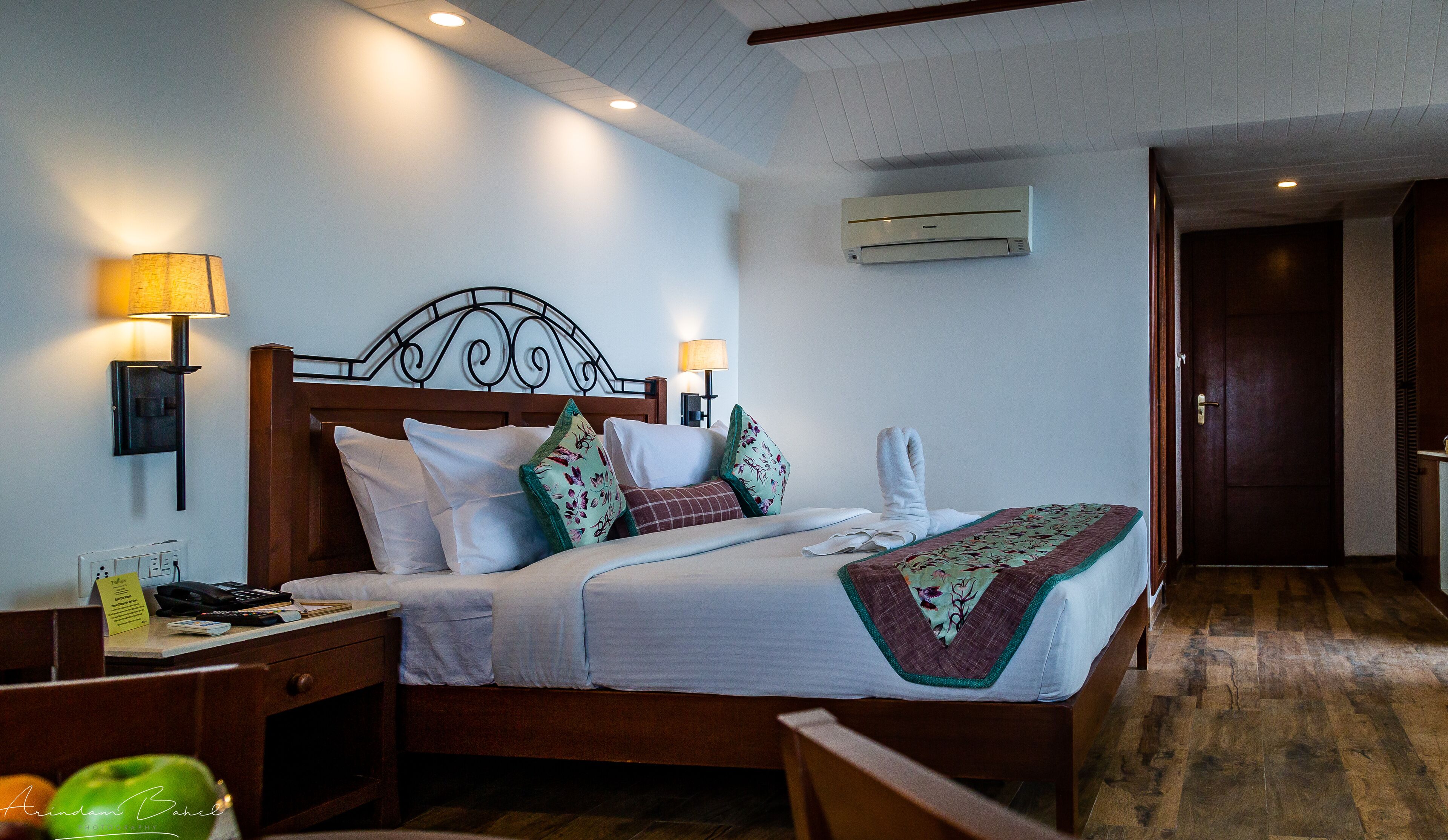 A hotel room with a wooden bed, white bedding, a nightstand with a lamp, a wall-mounted air conditioner, and a wooden floor.