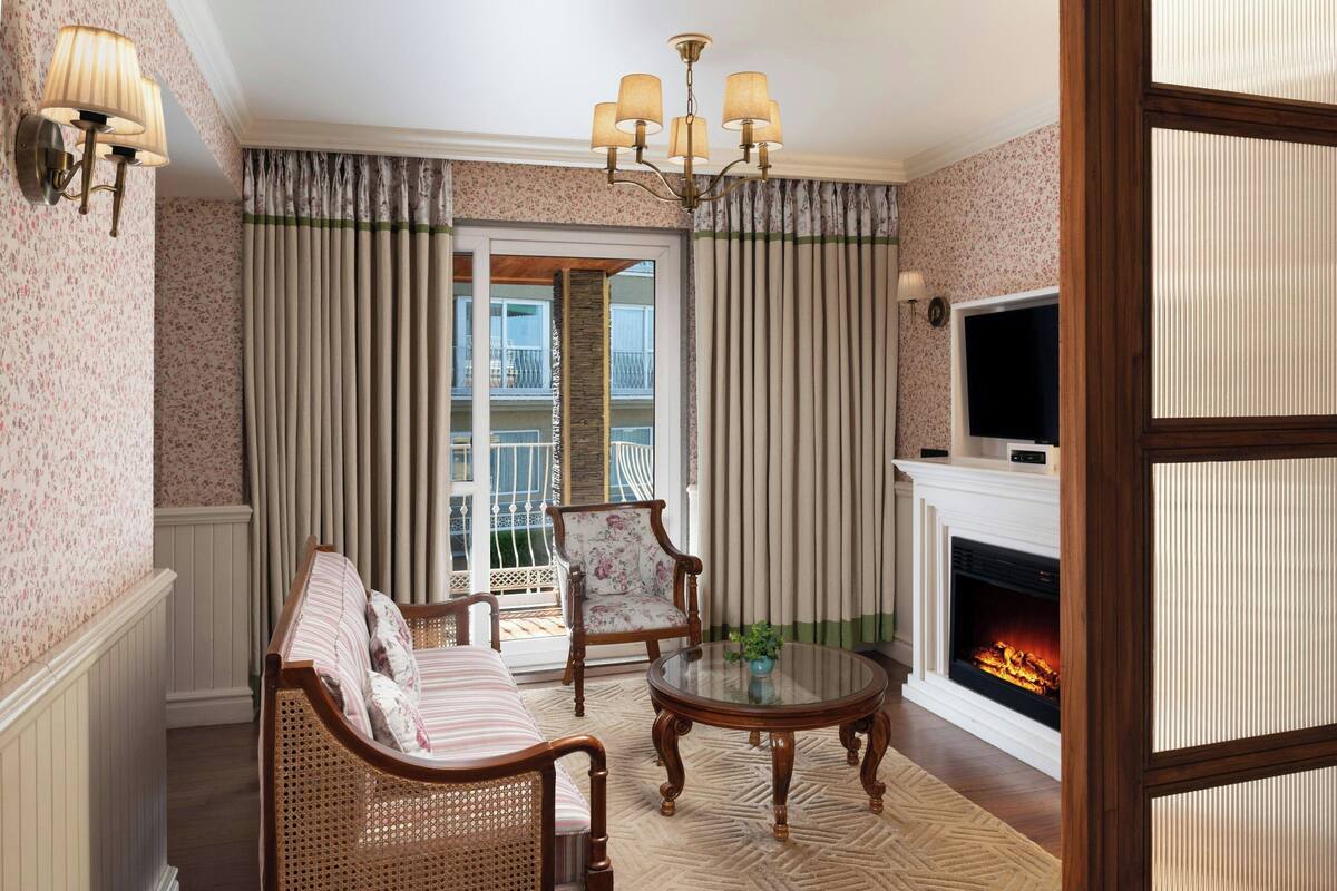 A cozy living room with a sofa, armchair, and a glass-top coffee table. A fireplace with a TV on the wall and a balcony with curtains are also visible.