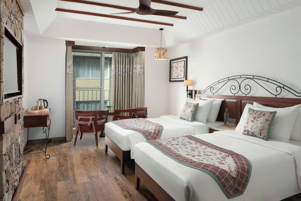 A hotel room with two beds, a wooden desk, a chair, a window with curtains, and a ceiling fan.