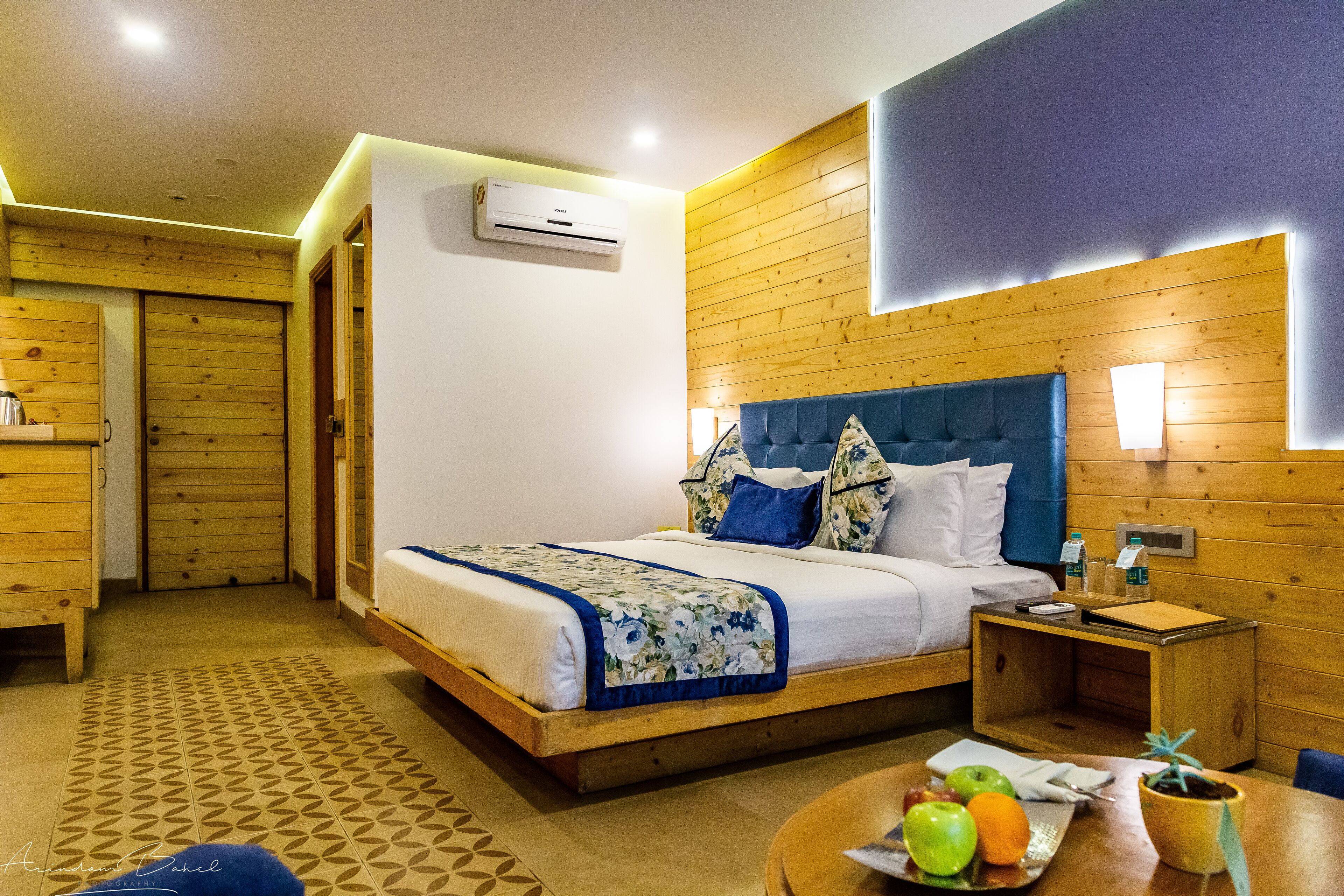 A hotel room with a wooden headboard, a bed with white and blue bedding, a bedside table with a lamp, and a small table with fruits and a plant.