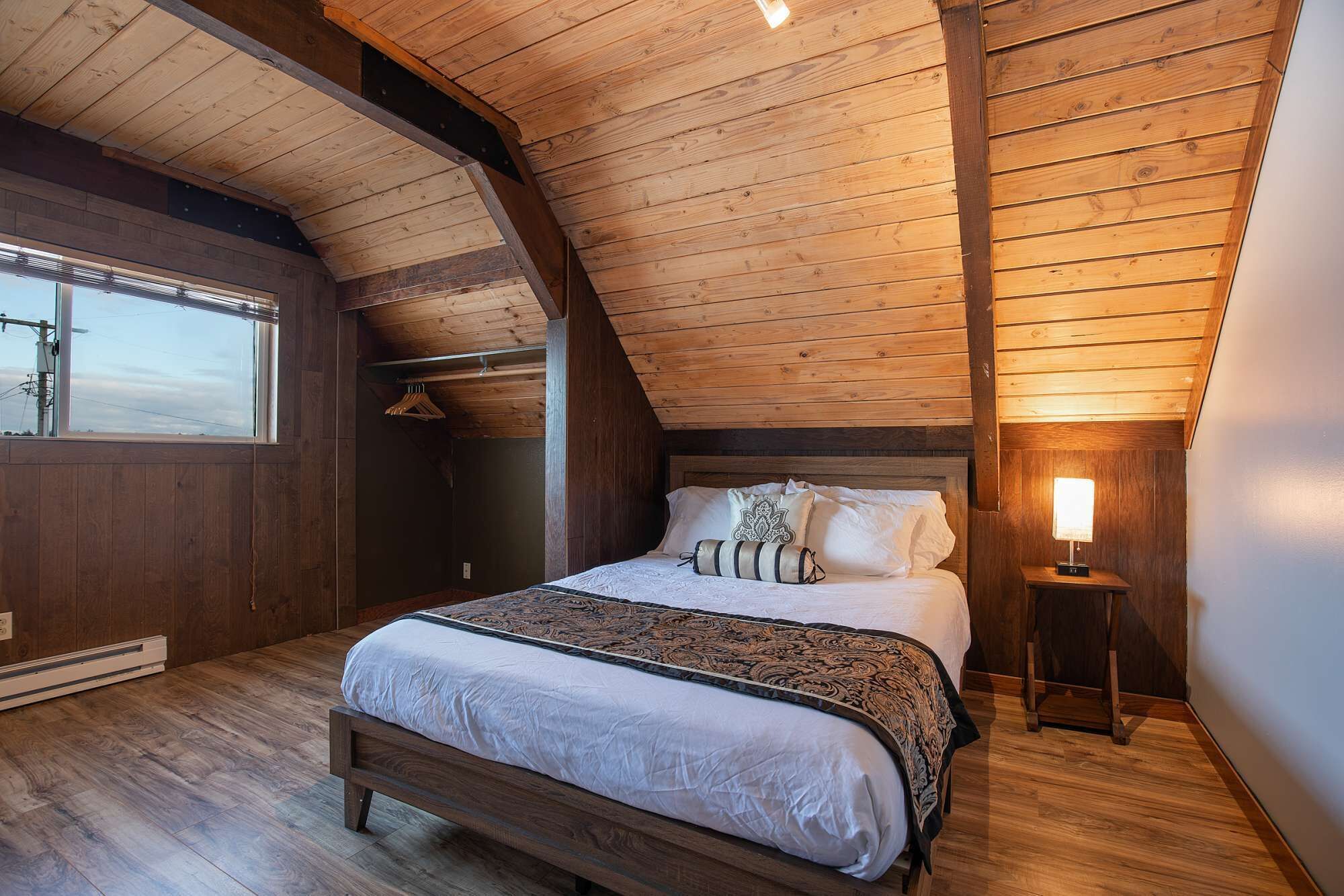 A bedroom with a sloped ceiling, a bed with patterned bedding, a bedside lamp, and a window with a view.