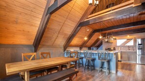 Ein geräumiges Loft mit hoher Decke, Holzbalken, einem Essbereich mit langem Tisch und Bänken sowie einer Küche mit modernen Geräten.