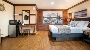A bedroom with a bed, a desk, a chair, a television, and a painting of a lake. - Flip Flop - Queen Studio Room at Wanderlust Inn, Pet Friendly (Ocean Shores)