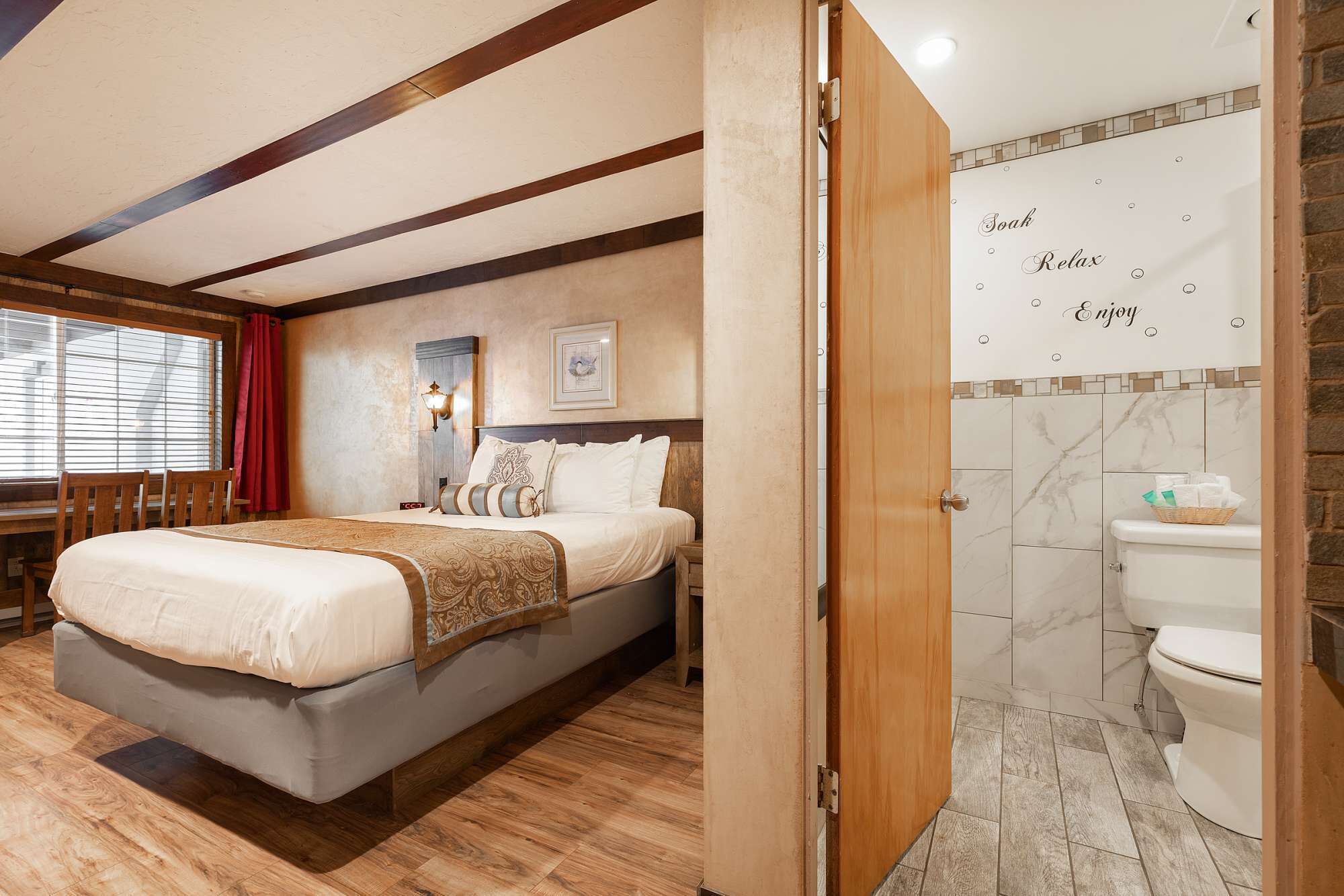 Habitación de hotel con una cama grande, un escritorio, una silla, una lámpara de pared, un cuadro enmarcado y un baño con inodoro y lavamanos.