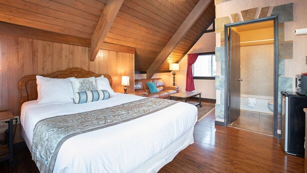 A spacious bedroom with a large bed, wooden ceiling beams, a sitting area with a sofa, and a view of the bathroom through an open door. - Ocean Song - Queen Studio Room at Wanderlust Inn, Pet Friendly (Ocean shores)