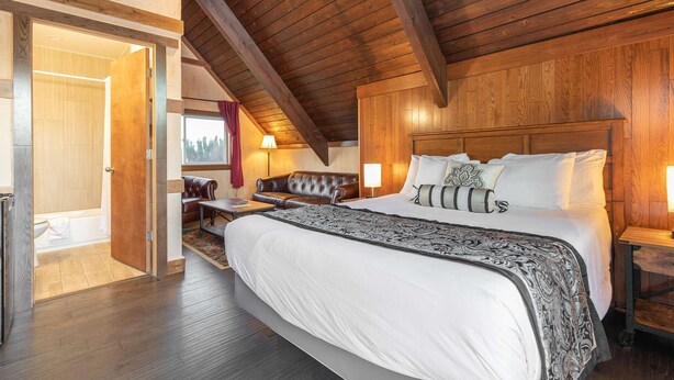 A spacious bedroom with a large bed, wooden ceiling beams, a seating area with a sofa, and a view of the outdoors through a window. - Once Upon a Tide - Queen Studio Room at Wanderlust Inn, Pet Friendly (Ocean shores)