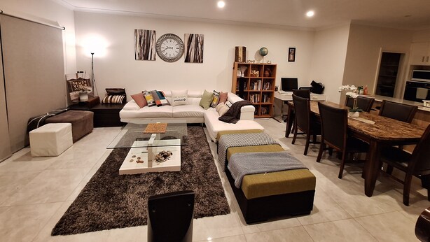 A modern living room with a sectional sofa, a coffee table, a bookshelf, and a dining area with a table and chairs. - New Serene 4 bedroom home (Kambah)