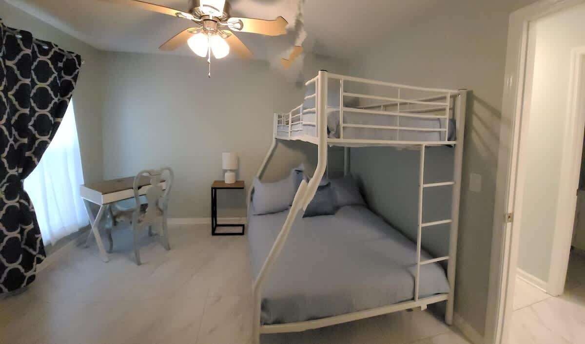 Une chambre avec un lit superposé, un bureau, une chaise, une petite table avec une lampe et un ventilateur de plafond.