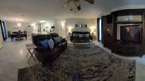A spacious living room with a large sofa, a coffee table, and a TV. There is a dining area with a table and chairs, and a kitchen in the background.