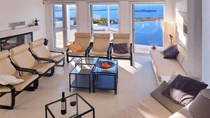 A modern living room with a sofa, chairs, a coffee table, and a TV. Large windows offer a view of the sea. - Luxury Private Villa with Infinity Pool & Sunset Sea Views & SPA (Paros)