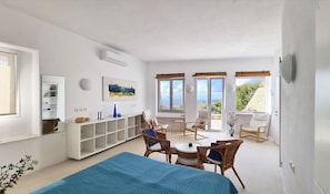 Une pièce spacieuse avec un grand lit, un coin repas avec des chaises en osier, une console blanche et une vue sur la mer grâce à de grandes fenêtres.