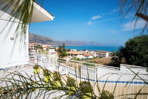 A balcony with potted plants, a white railing, and a view of a coastal town with buildings and mountains in the background. - VILLA TRINACRIA 1 PERFUMES OF SICILY (Alcamo marina)